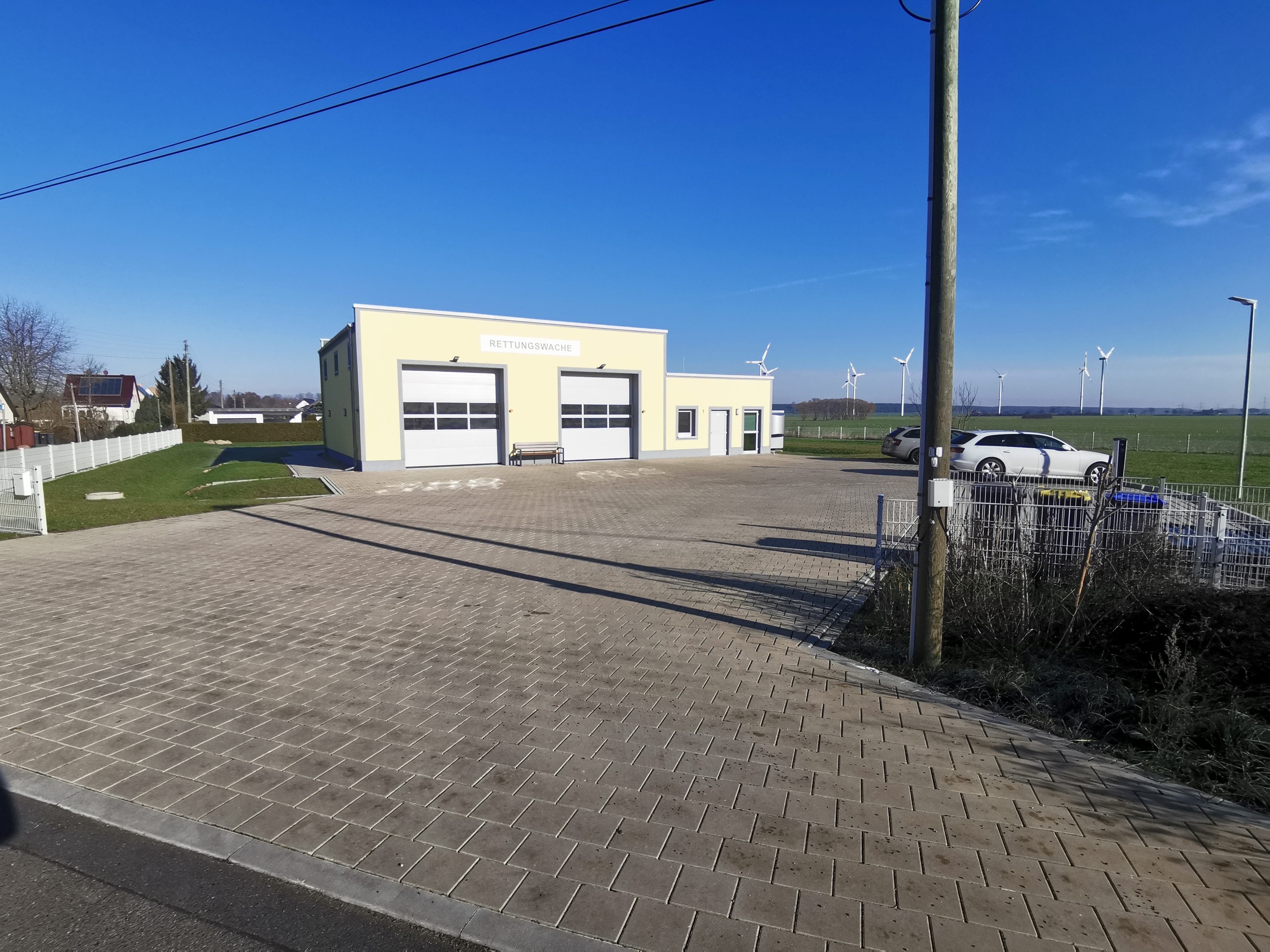 Einfahrt einer Rettungswache mit mehrseitigem Gefälle aus Betonpflastersteinen, quadratisch, mit Parkplatzanlage, Zaunanlage und Außenanlagenregulierung mit Rasenansaat